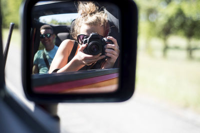 Woman photographing reflecting on side-view mirror while traveling with friend in off-road vehicle