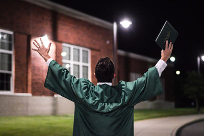 Rear view of man wearing graduation gown while standing against building at night