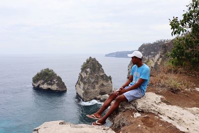 Man sitting on rock looking at sea against sky