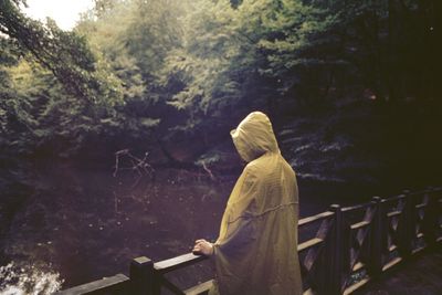Woman wearing raincoat looking at lake while standing on footbridge in forest