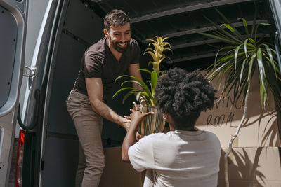Smiling boyfriend passing plant to multiracial girlfriend near van trunk