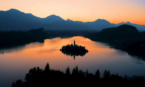 Scenic view of lake by silhouette mountains against orange sky