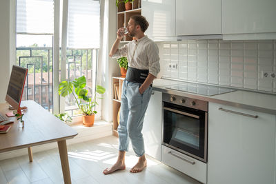 Man in back support corset on lower back to treatment of hernia on kitchen drinking water.