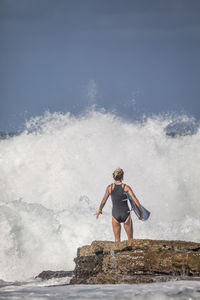 Rear view of woman with surfboard standing on rock at sea