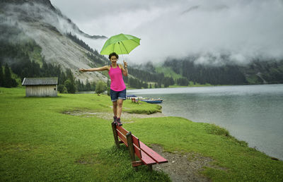 Playful woman holding umbrella while walking on bench at lakeshore against mountain range