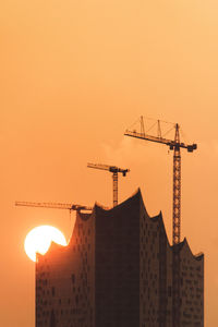 Silhouette crane by building against sky during sunset