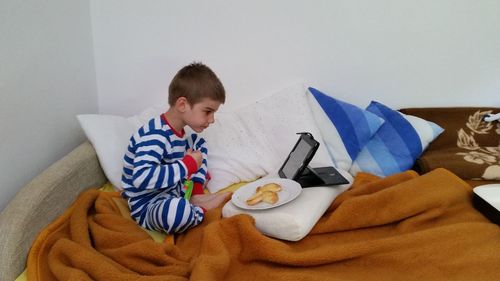 Boy watching on digital tablet while sitting by food in plate on bed