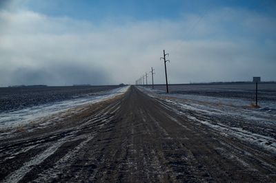 Dirt road alongside electricity pylons against sky