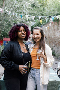 Young smiling female friends holding drinks while standing in back yard