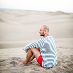 Side view of young man hugging knees while sitting at desert