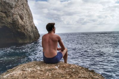 Rear view of shirtless man sitting on rock against sea at beach