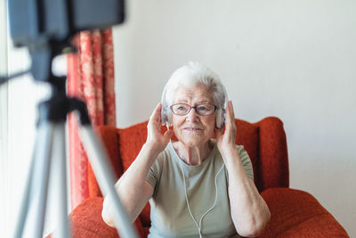 Content senior lady in headphones sitting in cozy armchair at home and enjoying songs while looking away