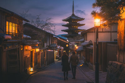 Rear view of woman walking on illuminated street amidst buildings at dusk