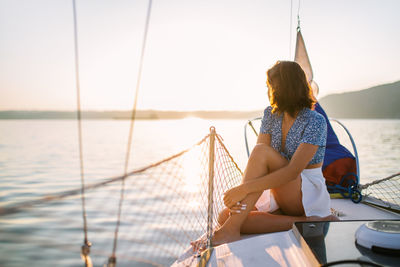 Side view of young stylish female traveler with dark hair in trendy crop top and skirt admiring sea while sitting on sailboat during cruise under sunset sky