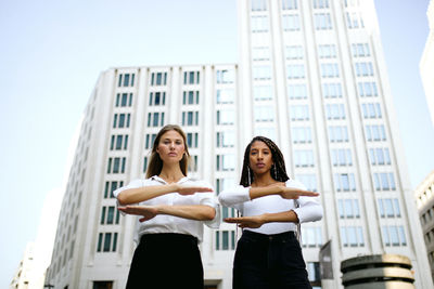Portrait of women gesturing equal sign while standing against buildings in city