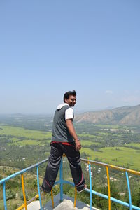 Rear view of man standing on railing against mountains and sky