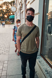 Young man walking along a store front in the city center in the evening wearing the face mask