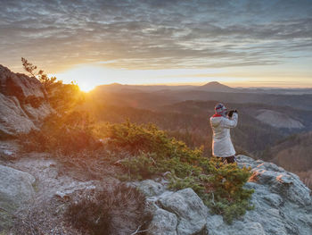 Photograper with hand on camera and tripod. woman climbed up on exposed rock for fall photos 