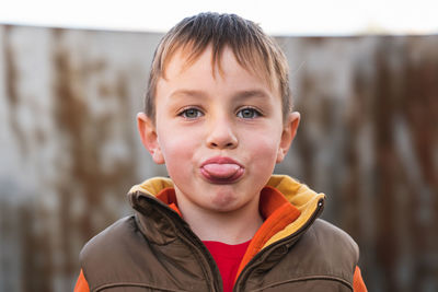 Close-up of a caucasian blond boy with green eyes wearing a brown fall vest showing his tongue.