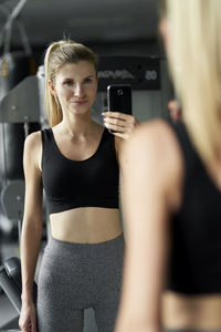 Portrait of woman taking selfie with mobile phone while looking into mirror at gym