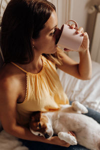 High angle view of young woman with dog drinking coffee in cup while sitting on bed