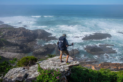 Rear view of man with hiking backpack and trekking pole looking at crashing waves onto sea rocks 