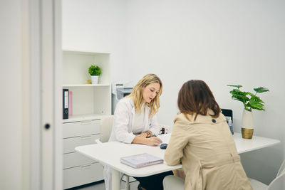 Serious female dermatologist sitting at table with client and taking notes on paper while writing prescription for buying cosmetic products