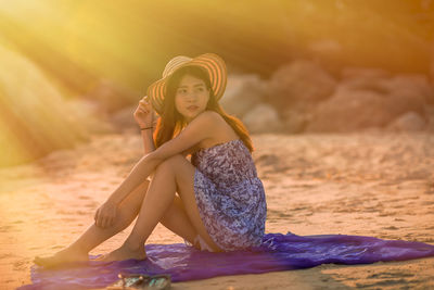 Portrait of young woman sitting on beach against sky during sunset