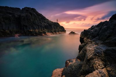 Distant view of man standing on cliff at shore against sky during sunset