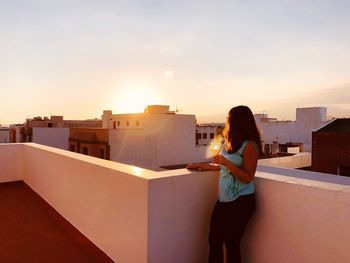 Woman standing on retaining wall against buildings in city at sunset