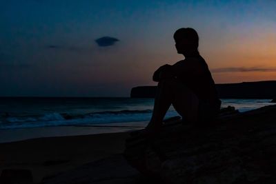 Silhouette man sitting on rock at beach against sky during sunset