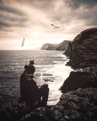 Man sitting on rock by sea against sky