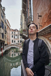Young man standing on canal amidst buildings in city