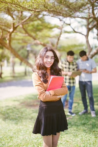 Portrait of smiling young woman with book standing at park
