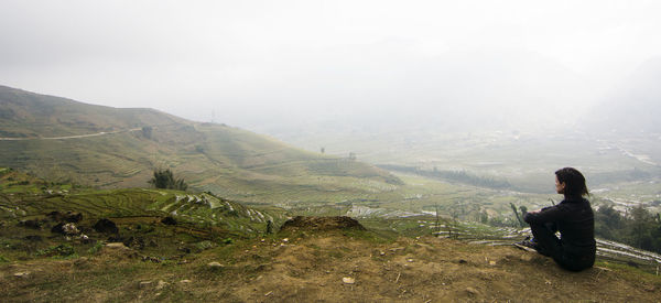 High angle view of woman sitting on cliff against rice terraces during foggy weather