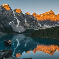 Rear view of friends standing on rock by lake against clear sky during sunset