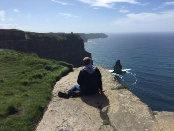 Rear view of man looking at sea while sitting on cliff against sky