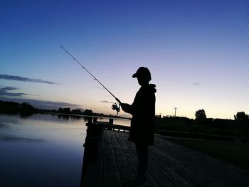 Side view of silhouette boy fishing on jetty over lake against sky during sunset
