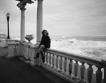 Portrait of woman sitting on railing by sea against sky