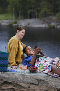 Female couple having picnic by river and using phone