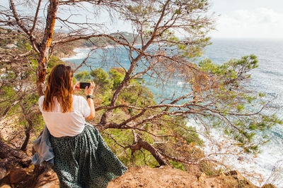 Woman photographing sea with smart phone while sitting on tree