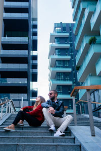 Low angle view of couple sitting on steps against buildings in city