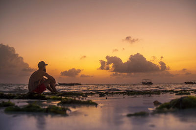 Man sitting on beach against sky during sunset