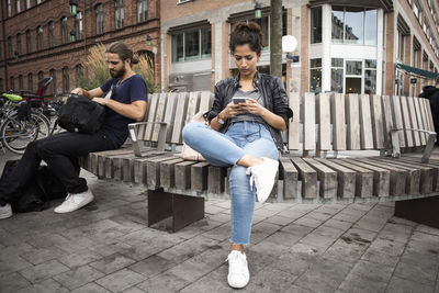 Low angle view of man and woman sitting on wooden bench against building in city