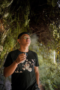 Young man looking away while standing against trees