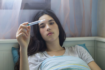 Close-up of teenage girl holding thermometer while lying on bed at home