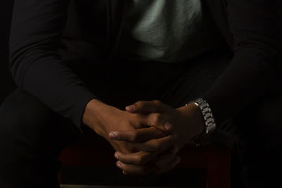 Midsection of man sitting with hands clasped against black background
