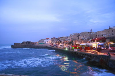 Illuminated buildings by sea against sky at dusk