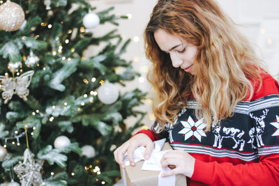 Pretty young girl opening presents near christmas tree. girl in red knitted sweater holds gift box.