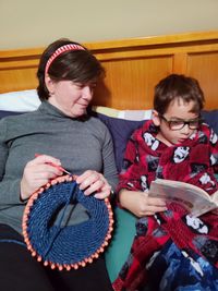 Mother looking at son reading book while sitting on bed
